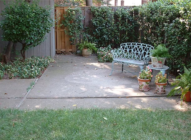 A charming backyard with a concrete patio, decorative metal bench, and lush greenery in a fenced gar.