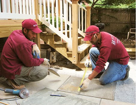 Two workers in maroon uniforms installing stone paving outdoors.