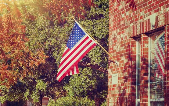 American flag hanging on brick building with autumn trees in background.